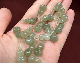 May include: A collection of small, pale green gourd-shaped charms. These charms are smooth and translucent, with a delicate, almost ethereal appearance. The charms are held in an open hand against a dark red background.