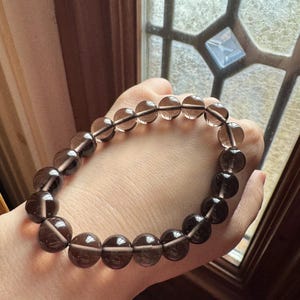 May include: A bracelet made of smoky quartz beads. The bracelet is composed of spherical, translucent beads in shades of brown and grey. The beads are strung together, forming a complete circle. The bracelet is displayed against a neutral background, highlighting the natural colour and texture of the stones.