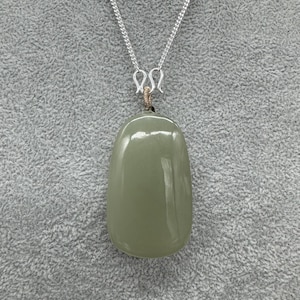 Puede incluir: Un colgante de piedra verde salvia, liso y pulido, con forma oblonga y redondeada. El colgante cuelga de una fina cadena de metal plateado mediante un peque&ntilde;o bucle de alambre enrollado que conecta la piedra con el cierre de metal plateado.