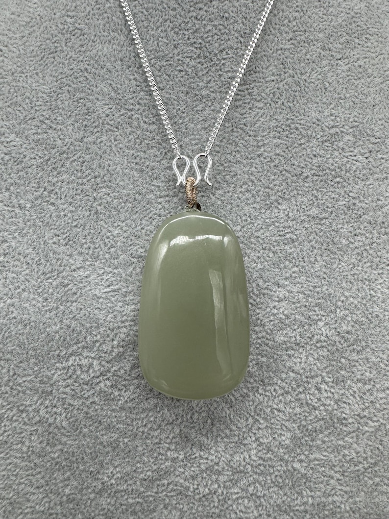 Puede incluir: Un colgante de piedra verde salvia, liso y pulido, con forma oblonga y redondeada. El colgante cuelga de una fina cadena de metal plateado mediante un peque&ntilde;o bucle de alambre enrollado que conecta la piedra con el cierre de metal plateado.