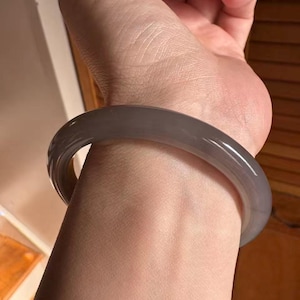 Könnte beinhalten: Ein graues Jade-Armreif-Armband. Das glatte, polierte Armband ist ein massives, abgerundetes Band. Das Armband wird am Handgelenk getragen, wobei die Hand und der Arm sichtbar sind. Der Jade hat eine durchscheinende Qualität.