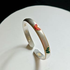 Pode incluir: Pulseira de punho prateada com acabamento escovado. A pulseira apresenta dois desenhos de borboletas, um vermelho e outro verde, com detalhes dourados. O design aberto facilita o uso.