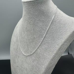 Puede incluir: Un collar de cadena fina de color plateado con un dise&ntilde;o cl&aacute;sico de eslabones tipo barbada. La cadena met&aacute;lica tiene un acabado pulido que refleja la luz, creando una apariencia brillante y uniforme mientras descansa sobre el busto.