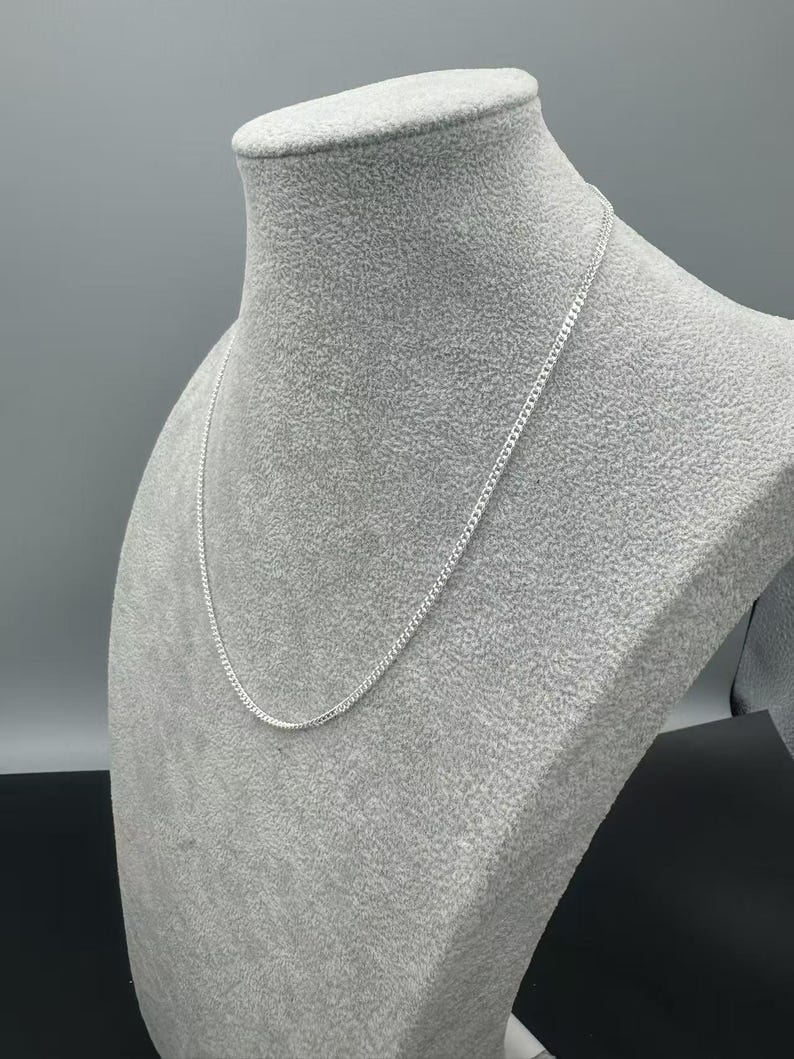 Puede incluir: Un collar de cadena fina de color plateado con un dise&ntilde;o cl&aacute;sico de eslabones tipo barbada. La cadena met&aacute;lica tiene un acabado pulido que refleja la luz, creando una apariencia brillante y uniforme mientras descansa sobre el busto.