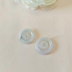 May include: A pair of circular, pale green jade pendants featuring a smooth, polished finish and a central hole. Each piece has a translucent quality with subtle natural veining and is attached to a small, clear plastic loop for hanging.