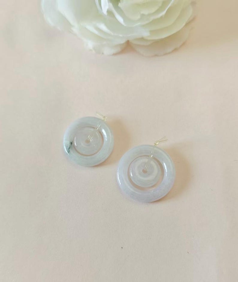 May include: A pair of circular, pale green jade pendants featuring a smooth, polished finish and a central hole. Each piece has a translucent quality with subtle natural veining and is attached to a small, clear plastic loop for hanging.