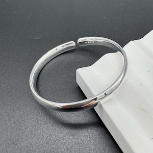 May include: A silver-colored, open-ended bangle bracelet. The bracelet has a smooth, polished surface and a simple, minimalist design. The bracelet is resting on a white surface against a dark gray background.