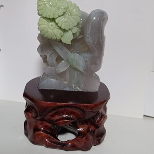 Può includere: Una scultura in giada di un'arrangiamento floreale su una base in legno. La giada è di colore verde pallido e i fiori sono scolpiti in uno stile realistico. La base è di colore marrone scuro e ha una texture naturale e ruvida.