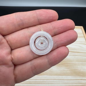 May include: A pale, translucent, off-white jade pendant featuring a double-ring design. The piece consists of a larger outer circular ring and a smaller inner circular ring, both connected by a thin, clear string loop at the top for suspension.