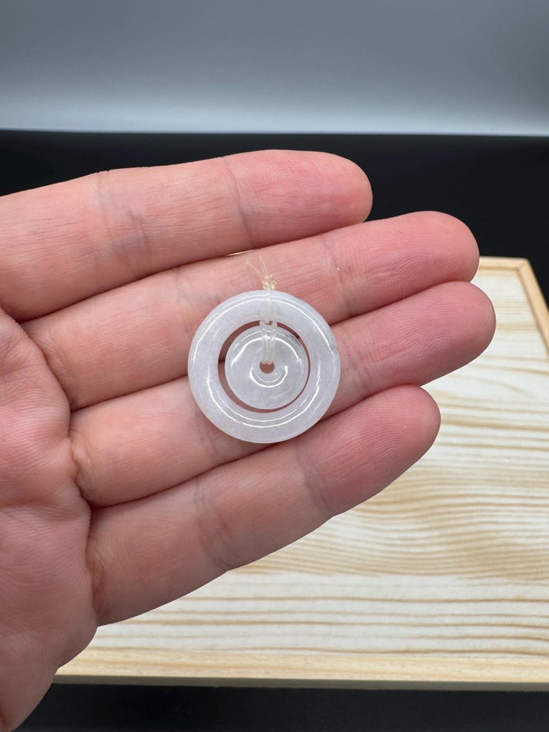 May include: A pale, translucent, off-white jade pendant featuring a double-ring design. The piece consists of a larger outer circular ring and a smaller inner circular ring, both connected by a thin, clear string loop at the top for suspension.