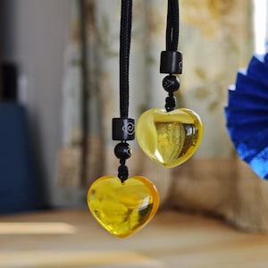May include: Two heart-shaped pendants with a yellow, translucent appearance hang from black cords. Each pendant has a black bead and a decorative black square. The background is blurred, with hints of a floral pattern and a blue paper fan.