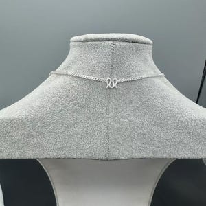 Puede incluir: Un collar de cadena de color plateado con un colgante central en forma de letra M. La cadena est&aacute; compuesta por peque&ntilde;os eslabones entrelazados que se extienden horizontalmente, mostrando un acabado met&aacute;lico y un dise&ntilde;o sencillo.