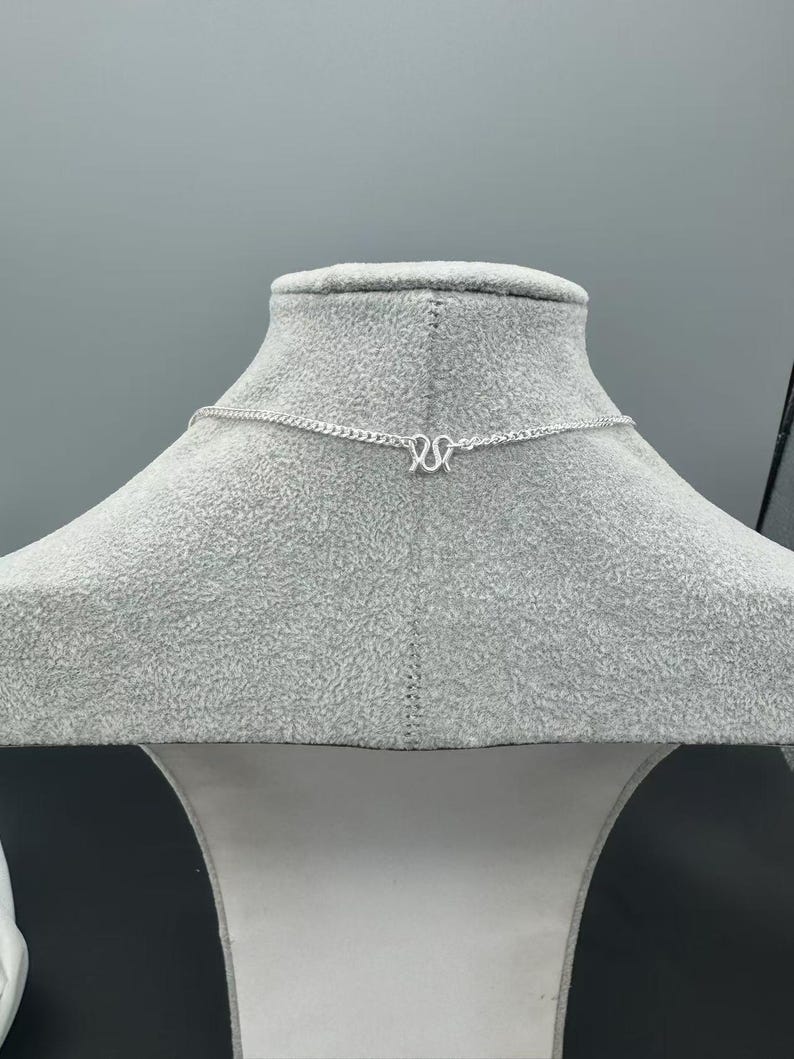 Puede incluir: Un collar de cadena de color plateado con un colgante central en forma de letra M. La cadena est&aacute; compuesta por peque&ntilde;os eslabones entrelazados que se extienden horizontalmente, mostrando un acabado met&aacute;lico y un dise&ntilde;o sencillo.