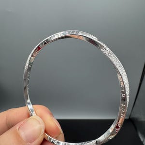 Pode incluir: Uma pulseira de bracelete prateada com uma faixa texturizada e brilhante. A pulseira tem o texto gravado "time does not disperse we do not disperse". A pulseira é segurada em uma mão contra um fundo cinza.