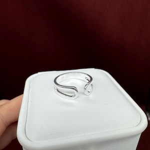 May include: A silver-coloured open ring featuring a smooth, polished finish and a continuous loop design that creates a gap at the front. The band has a rounded profile and a reflective metallic surface, showcasing a simple and clean aesthetic.