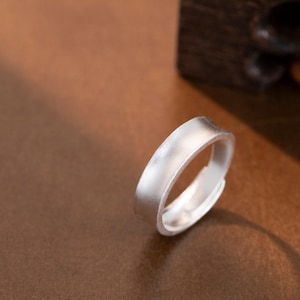 May include: A silver ring with a flat, brushed metal band. The ring is adjustable and rests on a brown surface. The ring's design is simple and modern, suitable for everyday wear.