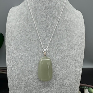 Puede incluir: Un colgante de piedra lisa de forma ovalada en color verde salvia, suspendido de una fina cadena de plata. El colgante est&aacute; unido a la cadena mediante una peque&ntilde;a anilla de alambre enrollado, con una superficie pulida y un tono verde suave.