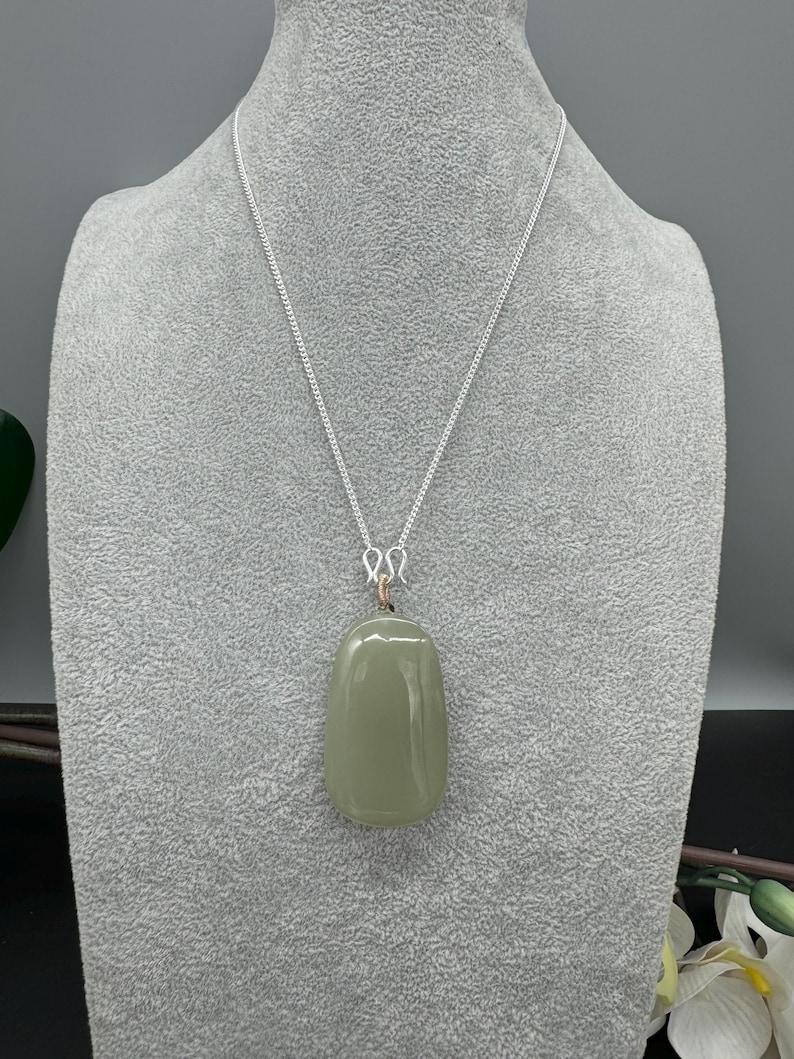 Puede incluir: Un colgante de piedra lisa de forma ovalada en color verde salvia, suspendido de una fina cadena de plata. El colgante est&aacute; unido a la cadena mediante una peque&ntilde;a anilla de alambre enrollado, con una superficie pulida y un tono verde suave.