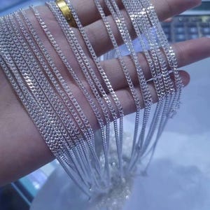 Può includere: Una collezione di molteplici catene a maglia barbazzale di colore argento con maglie piatte intrecciate. Le catene metalliche sono drappeggiate sul palmo, mostrando una finitura lucida e riflettente e un motivo a maglie coerente.