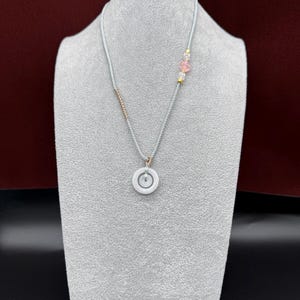May include: A necklace featuring a light gray cord with a central white circular pendant. The cord is accented with a small section of metallic beads on one side and a cluster of pink and clear translucent beads with gold spacers on the other side.