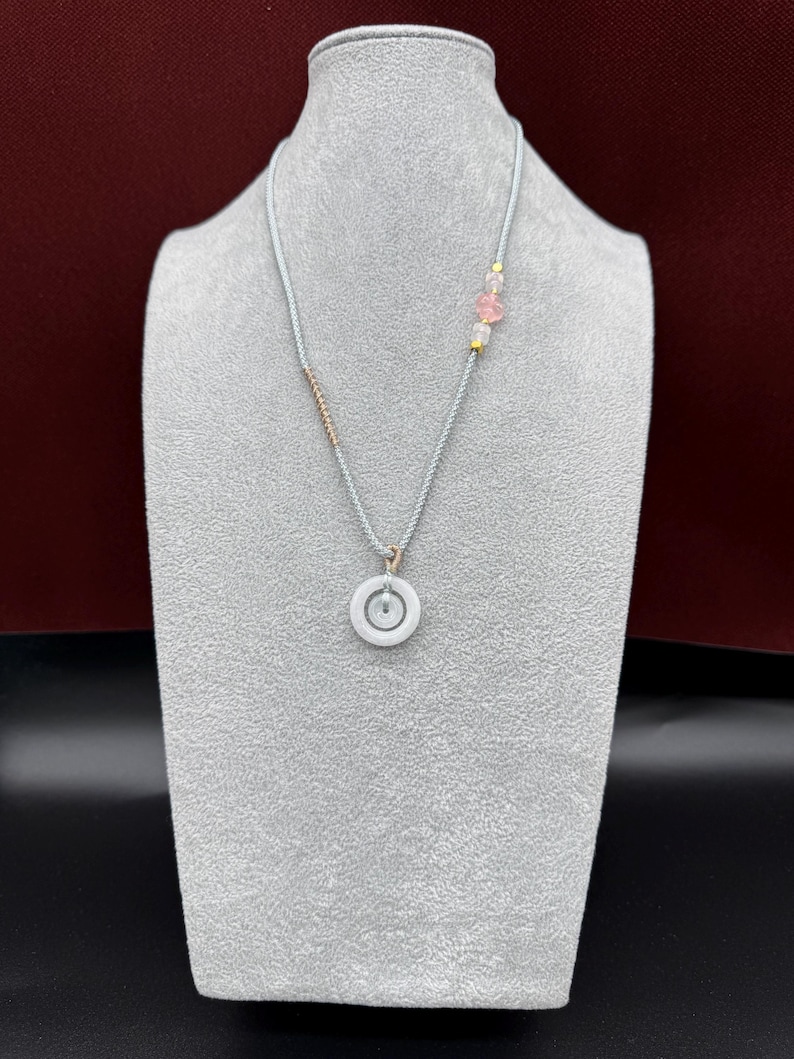 May include: A necklace featuring a light gray cord with a central white circular pendant. The cord is accented with a small section of metallic beads on one side and a cluster of pink and clear translucent beads with gold spacers on the other side.