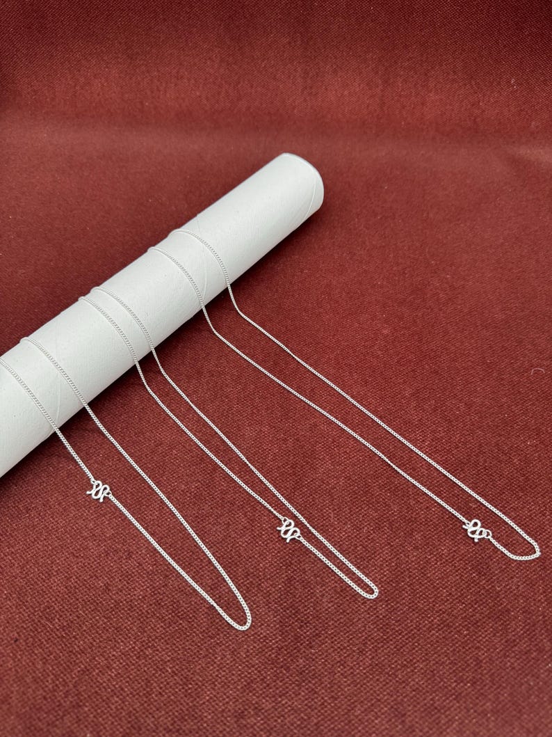 Puede incluir: Tres cadenas de metal de color plateado, cada una con un peque&ntilde;o y detallado colgante en forma de letra cursiva. Las delicadas cadenas de eslabones est&aacute;n colocadas sobre un tubo de exhibici&oacute;n cil&iacute;ndrico blanco, resaltando el acabado met&aacute;lico.