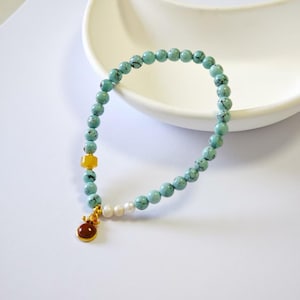 Peut inclure: Un bracelet de perles turquoise avec un charm doré et une perle jaune. Le bracelet comporte également des perles blanches. Le charm est une petite pierre ronde brun-rougeâtre avec des accents dorés. Le bracelet est sur une surface blanche.
