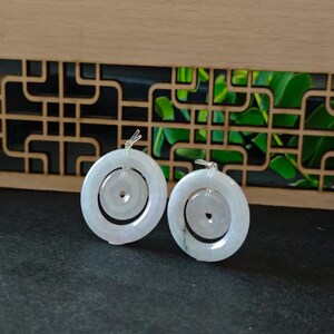 May include: A pair of pale, translucent white jade pendants, each featuring a smaller circular disc suspended within a larger circular ring. The smooth, polished stone pieces are connected by small, clear plastic loops at the top of each ring.