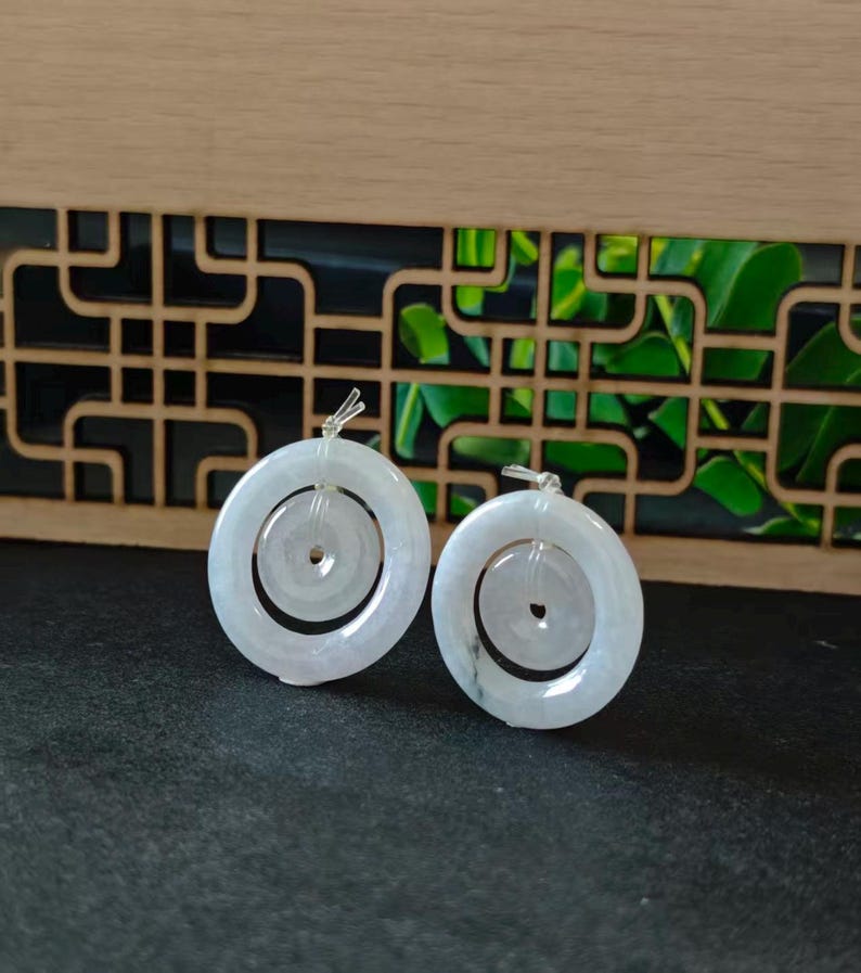 May include: A pair of pale, translucent white jade pendants, each featuring a smaller circular disc suspended within a larger circular ring. The smooth, polished stone pieces are connected by small, clear plastic loops at the top of each ring.