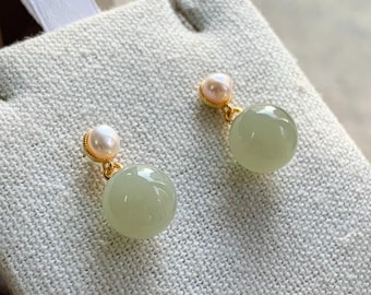 May include: A pair of elegant earrings featuring a light green, spherical jade stone suspended from a small, pink pearl. The earrings have gold-colored accents and are displayed on a textured, off-white surface.