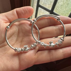 Pode incluir: Duas pulseiras de bebê de prata com contas decorativas e pingentes em forma de coroa. As pulseiras ajustáveis são seguradas em uma mão, mostrando o metal brilhante. Estas pulseiras são um presente tradicional para bebês.