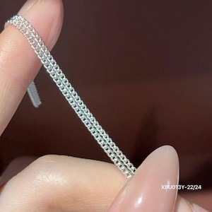 Puede incluir: Un collar de cadena de plata de doble hebra con eslabones redondos peque&ntilde;os, uniformes y entrelazados. El metal tiene un acabado brillante y reflectante. El texto XBU013Y-22/24 est&aacute; impreso en blanco en la parte inferior derecha de los eslabones.