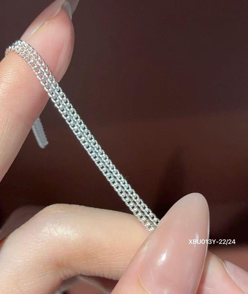 Puede incluir: Un collar de cadena de plata de doble hebra con eslabones redondos peque&ntilde;os, uniformes y entrelazados. El metal tiene un acabado brillante y reflectante. El texto XBU013Y-22/24 est&aacute; impreso en blanco en la parte inferior derecha de los eslabones.