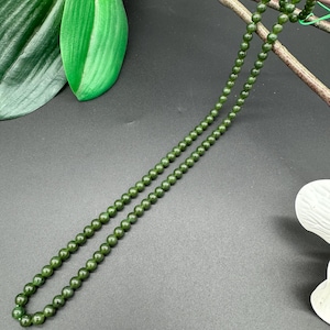 Puede incluir: Un collar largo de cuentas verde oscuro. El collar está compuesto por pequeñas cuentas redondas. El fondo es una superficie gris oscuro con una hoja verde y una flor blanca en la esquina superior derecha.