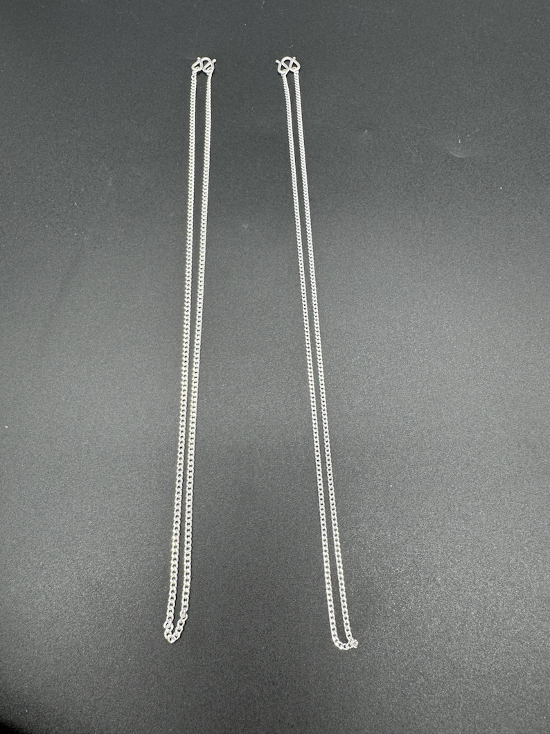 Puede incluir: Dos cadenas de metal de color plateado id&eacute;nticas, cada una con un dise&ntilde;o de eslabones finos tipo barbada. Ambas cadenas son largas y delgadas, est&aacute;n suspendidas verticalmente y terminan con un peque&ntilde;o mecanismo de cierre seguro en la parte superior.