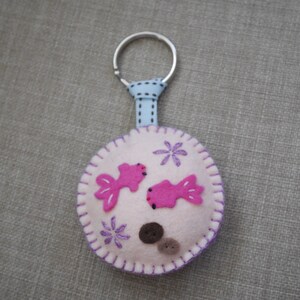 May include: A pink felt keychain with two pink fish and purple flowers. The keychain is attached to a silver ring and a blue ribbon.
