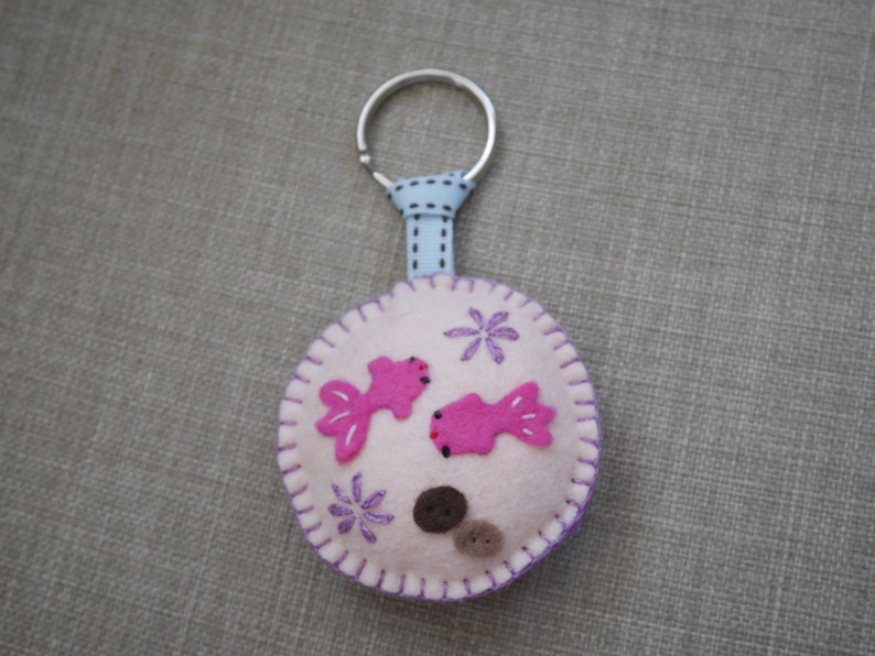May include: A pink felt keychain with two pink fish and purple flowers. The keychain is attached to a silver ring and a blue ribbon.