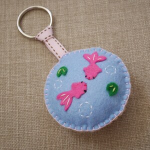May include: A blue felt keychain with a pink fish design and green leaves. The keychain has a silver metal ring.