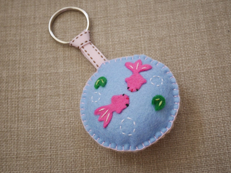May include: A blue felt keychain with a pink fish design and green leaves. The keychain has a silver metal ring.