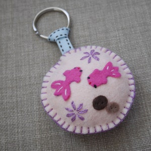May include: A white felt keychain with a pink fish design, purple embroidered flowers, and brown felt dots. The keychain has a silver metal ring and a blue ribbon.