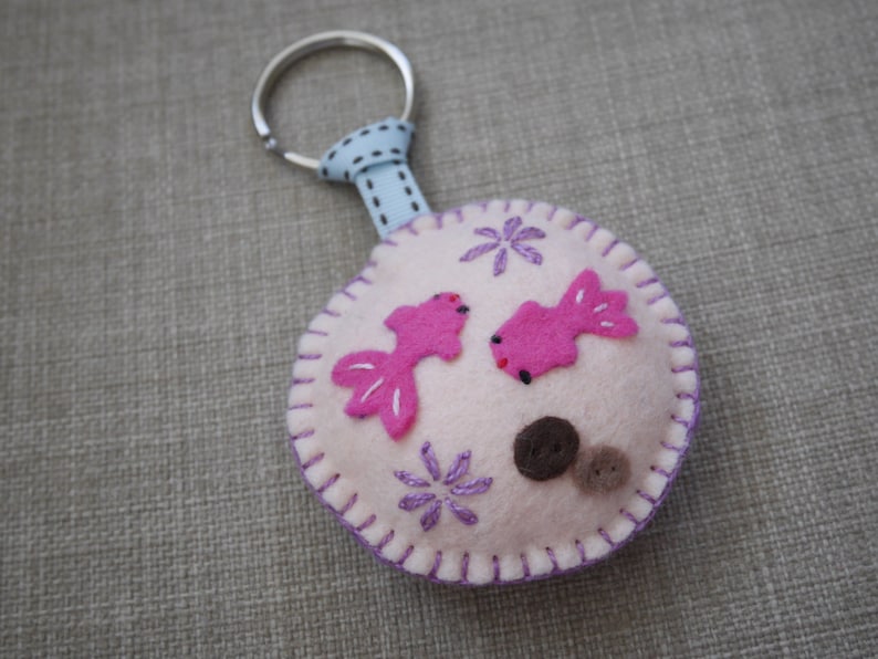 May include: A white felt keychain with a pink fish design, purple embroidered flowers, and brown felt dots. The keychain has a silver metal ring and a blue ribbon.
