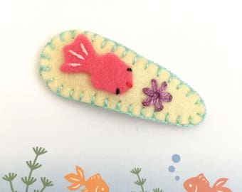 May include: A yellow felt hair clip with a pink felt fish and a purple felt flower. The fish has white felt fins and black embroidered eyes.