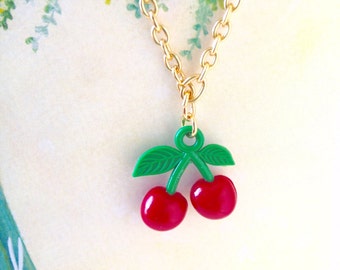 May include: A gold chain necklace with a red and green cherry charm. The cherry charm has a green stem and leaves.