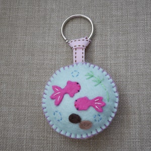 May include: A light blue felt keychain with pink fish and brown rocks, attached to a silver key ring with a pink ribbon.