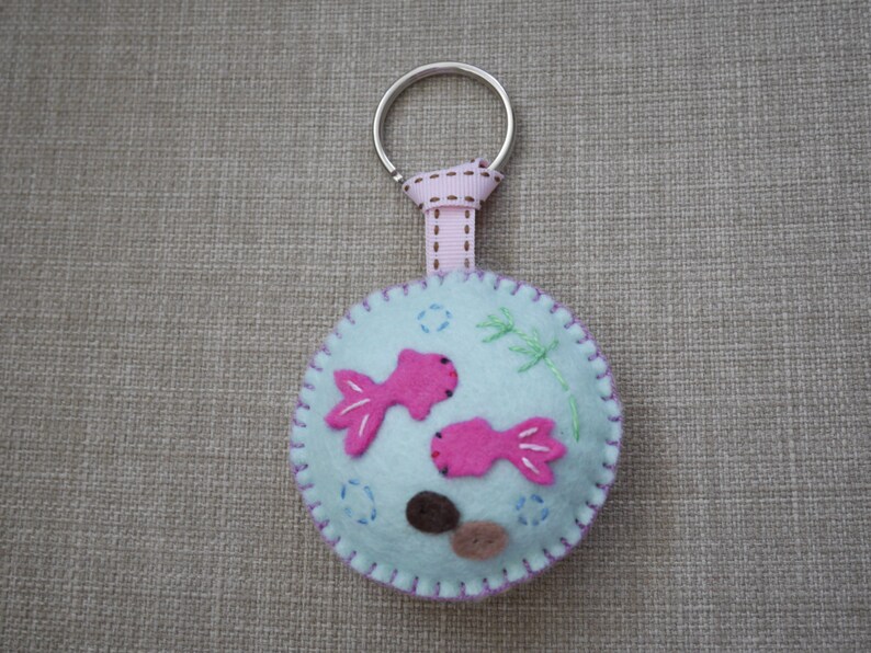 May include: A light blue felt keychain with pink fish and brown rocks, attached to a silver key ring with a pink ribbon.