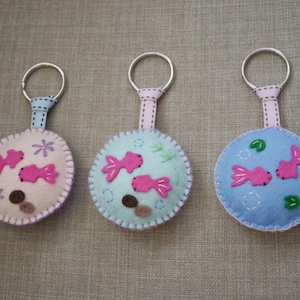 May include: Three felt keychains with pink fish and blue water designs. Each keychain has a silver ring and a pink ribbon.