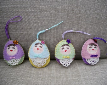 May include: Four handmade felt doll ornaments with embroidered faces and lace trim. Each doll is wearing a different color dress: purple, light blue, green, and pink.