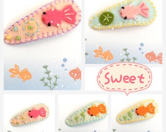 May include: A collection of handmade felt hair clips featuring goldfish designs. Each clip has a unique color scheme, including pink, orange, and yellow fish, with embroidered details and decorative stitching. The word "Sweet" is in a pink speech bubble.