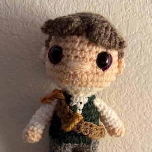 May include: A handmade crochet doll of a person with brown hair, large black eyes, and a tan complexion. The doll wears a white shirt, a dark green vest, gray pants, and brown boots. A brown satchel is across the chest.