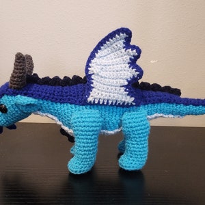 May include: A handmade crochet dragon in shades of blue, with a white and blue wing design. The dragon has dark grey horns and black eyes. The crochet toy is standing on a dark surface.